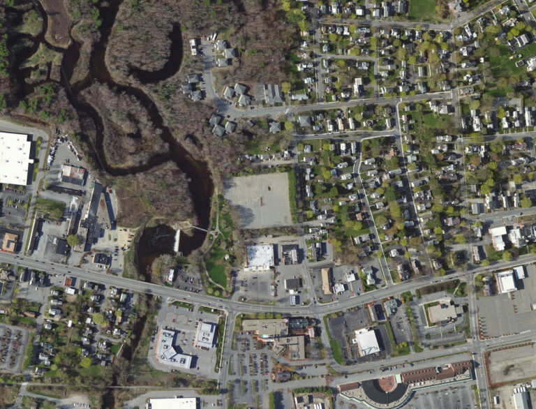 Aerial view of a small New England city, with a rectangular vacant lot at center surrounded by the winding Ashuelot River and its wetlands, residential neighborhoods, and commercial development.