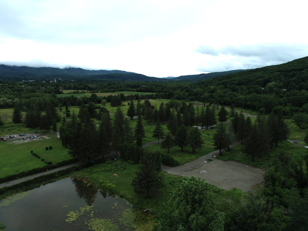 Aerial drone photograph of Spruces Park in Williamstown, Massachusetts, showing a broad, flat floodplain landscape with scattered mature evergreen trees, open green lawns, a pond with aquatic vegetation in the foreground, and a bare gravel area to the right. Tree-lined hills and low mountains recede into a cloudy sky in the background, with the village of Williamstown visible at the upper left.