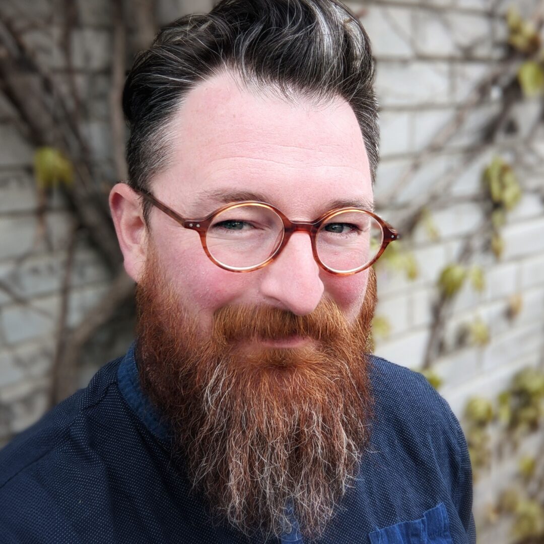 Nate Card's headshot: bearded person wearing round tortoiseshell glasses and a dark blue shirt