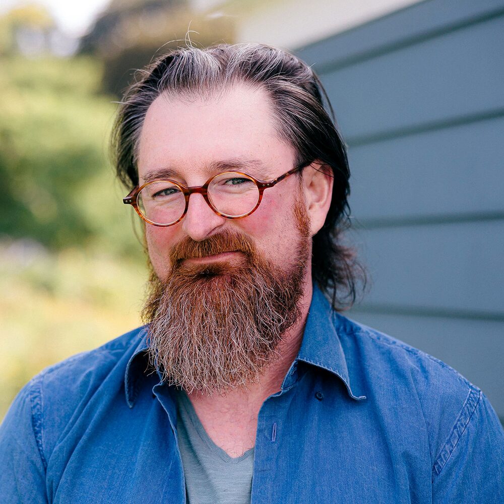 Nate Card's headshot: bearded person wearing round tortoiseshell glasses and a dark blue shirt