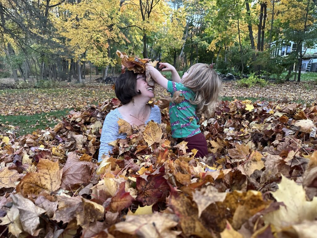 An adult and a young child laugh together while playing in a large pile of fallen maple leaves, surrounded by golden autumn trees.