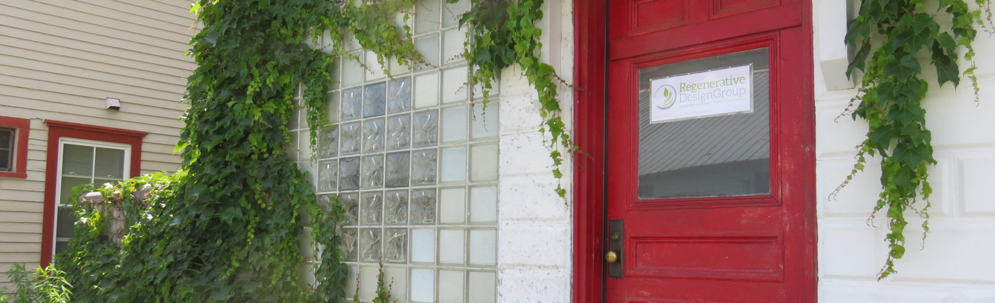A white brick building with climbing vines and a red door, surrounded by plants