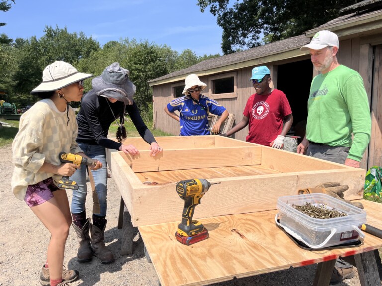Five people work together outdoors to assemble a wooden air-prune nursery bed, using power drills and screws, with farm buildings visible in the background.