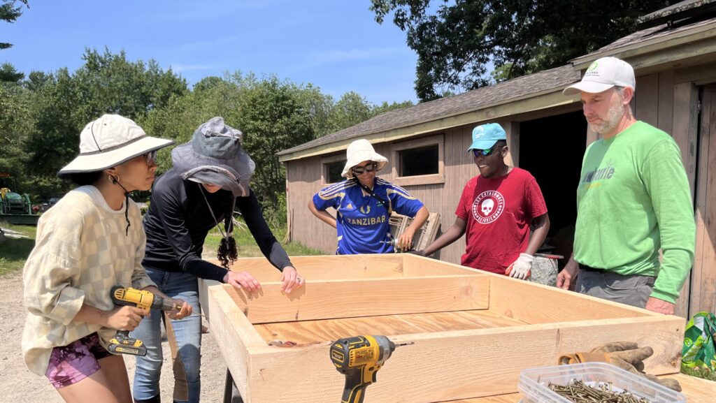 Five people work together outdoors to assemble a wooden air-prune nursery bed, using power drills and screws, with farm buildings visible in the background.