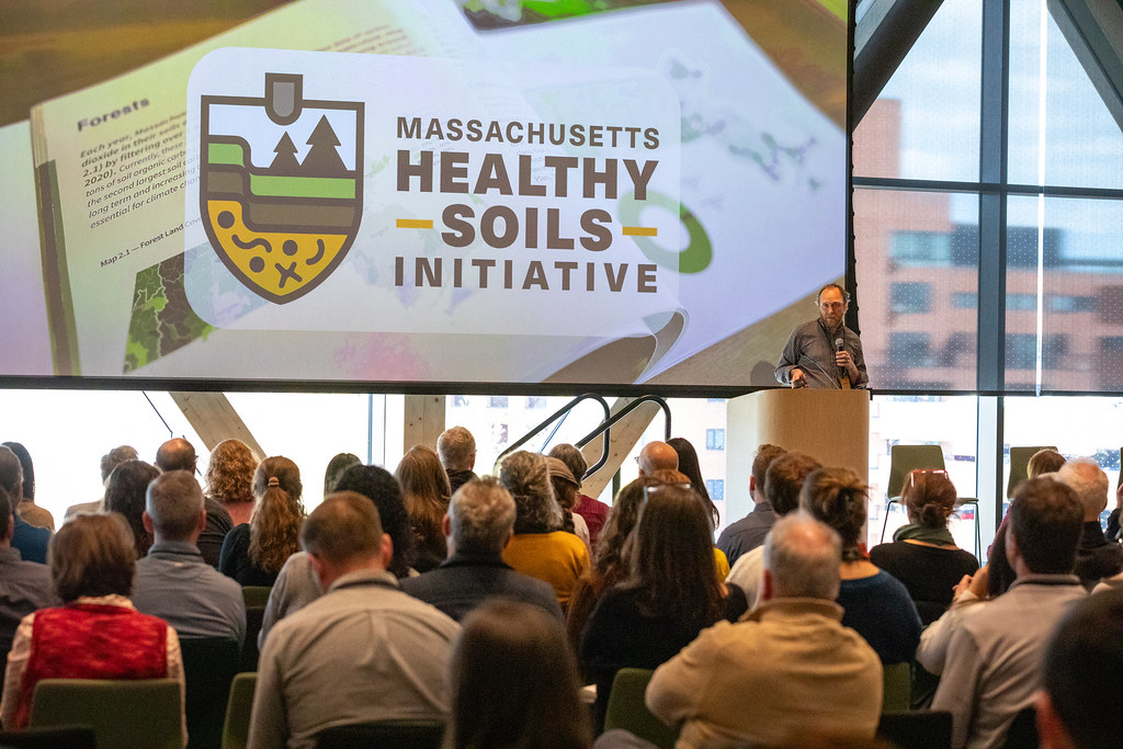 A speaker stands at a podium at the front of a large auditorium-style room with floor-to-ceiling windows. A large projected slide behind him displays the Massachusetts Healthy Soils Initiative logo over a background image of maps and soil layers. A full audience is visible from behind in the foreground.
