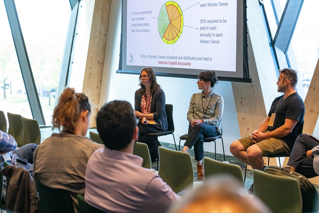 Three panelists seated in chairs in a bright wood-paneled room, with a projected slide showing a pie chart diagram of worker-owner dividend distribution. Two audience members are visible in the foreground with their backs to the camera.