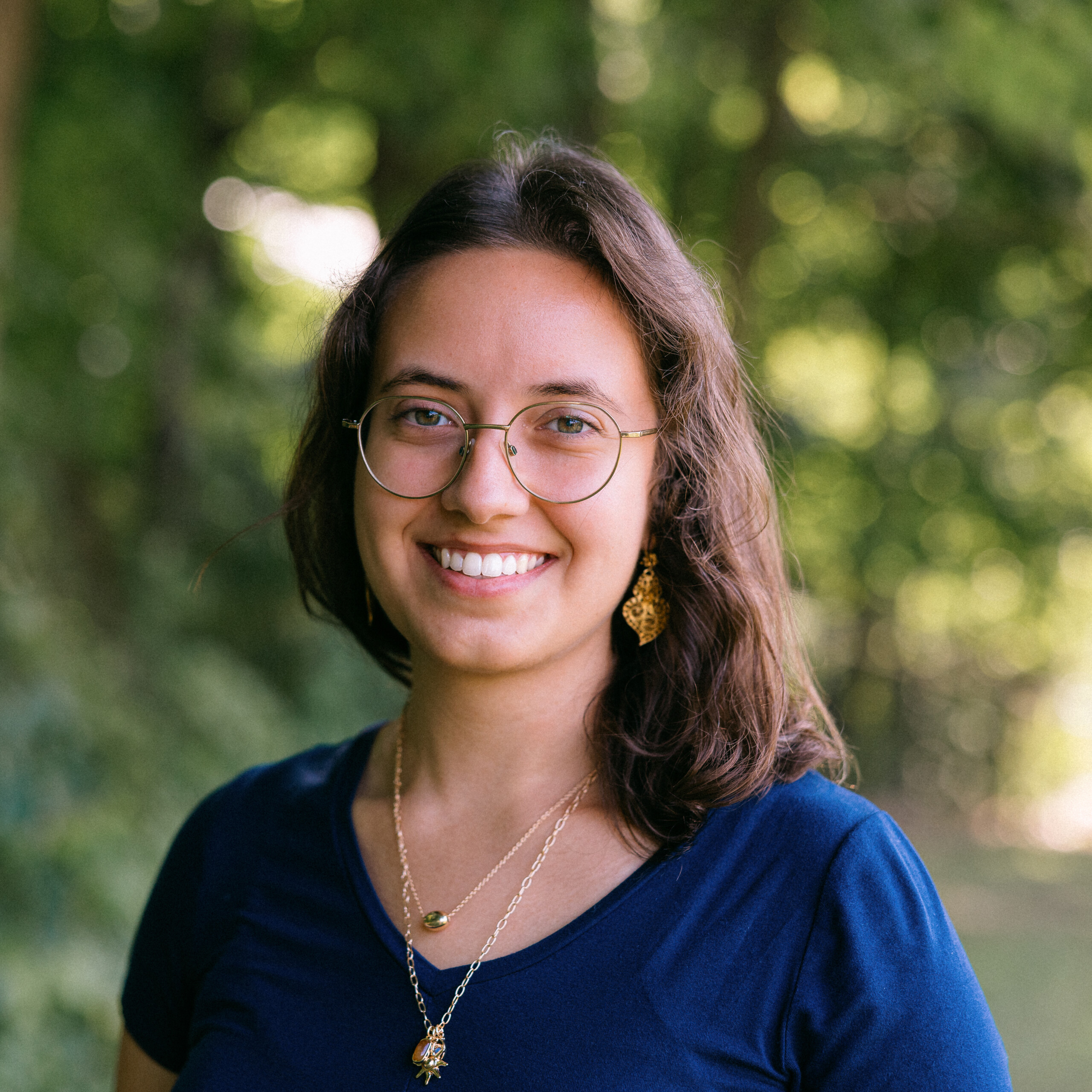 Daniella Portal, a woman with mid-length brown hair, wearing glasses and two pendant necklaces, as well as a deep blue shirt. She is smiling at the camera. A blurred nature scene in the background.