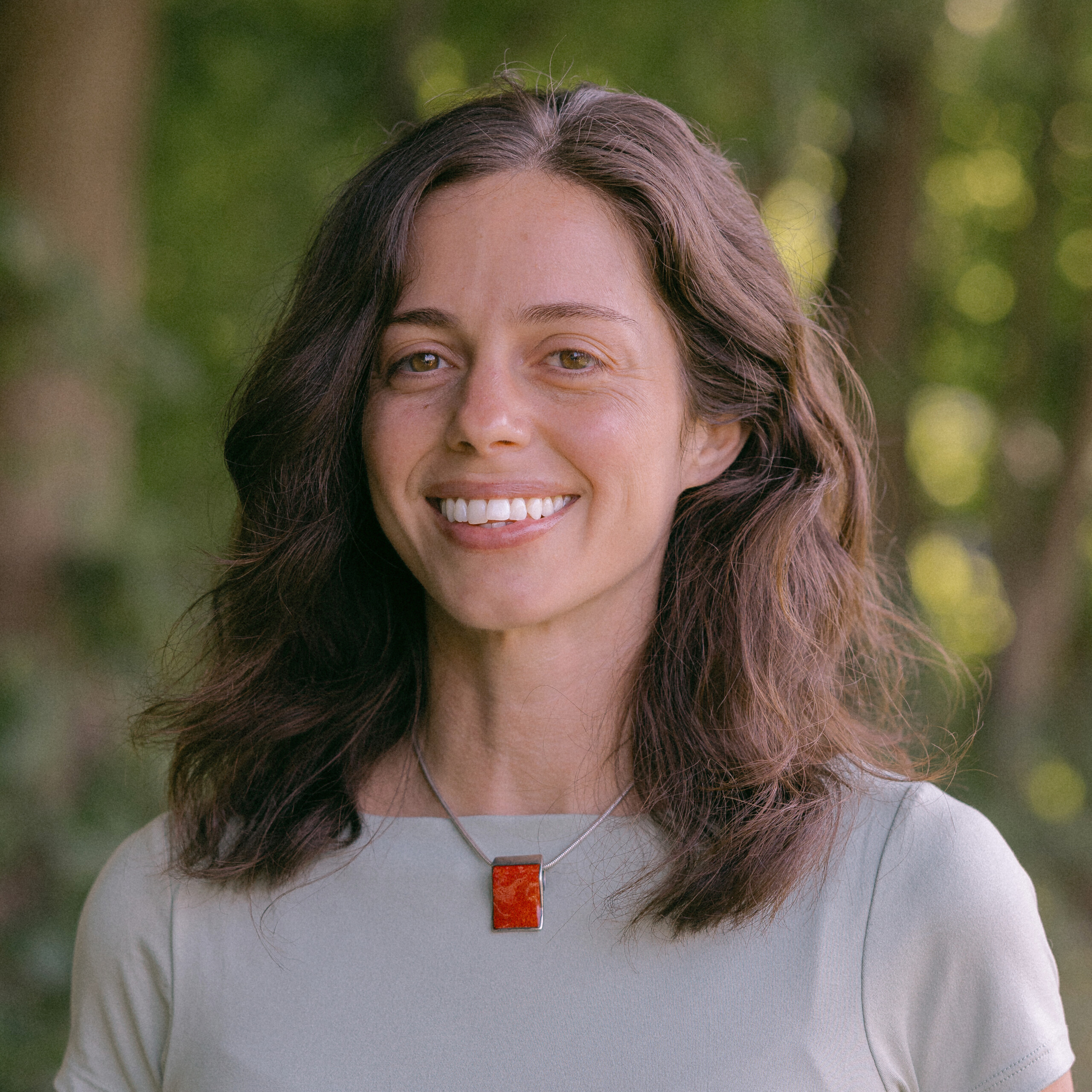 Maria Biello - a woman with brown hair and wearing a gray shirt and red square pendant necklace, smiling at the camera with a blurred nature background